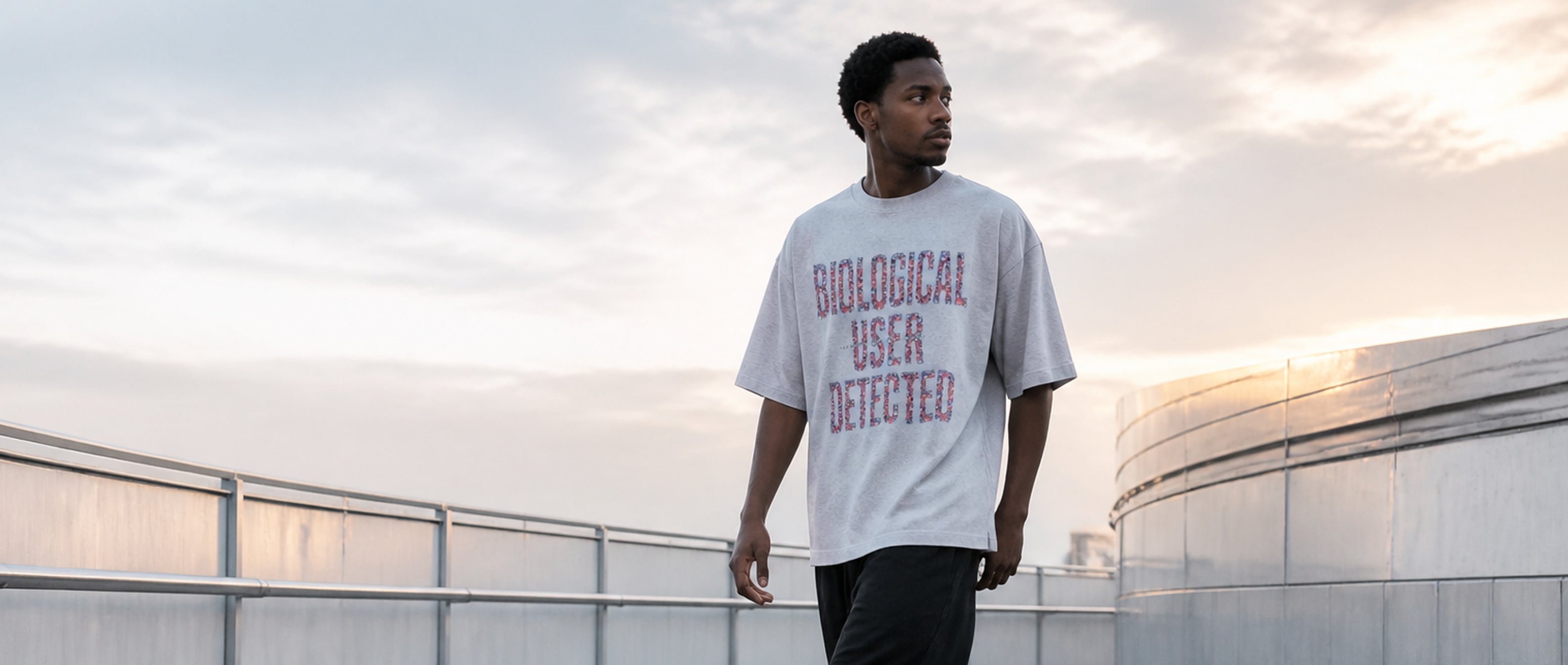 Adult model wearing the 3o1d BIOLOGICAL USER DETECTED oversized T-shirt in Cool Heather Grey on a sunrise overpass.